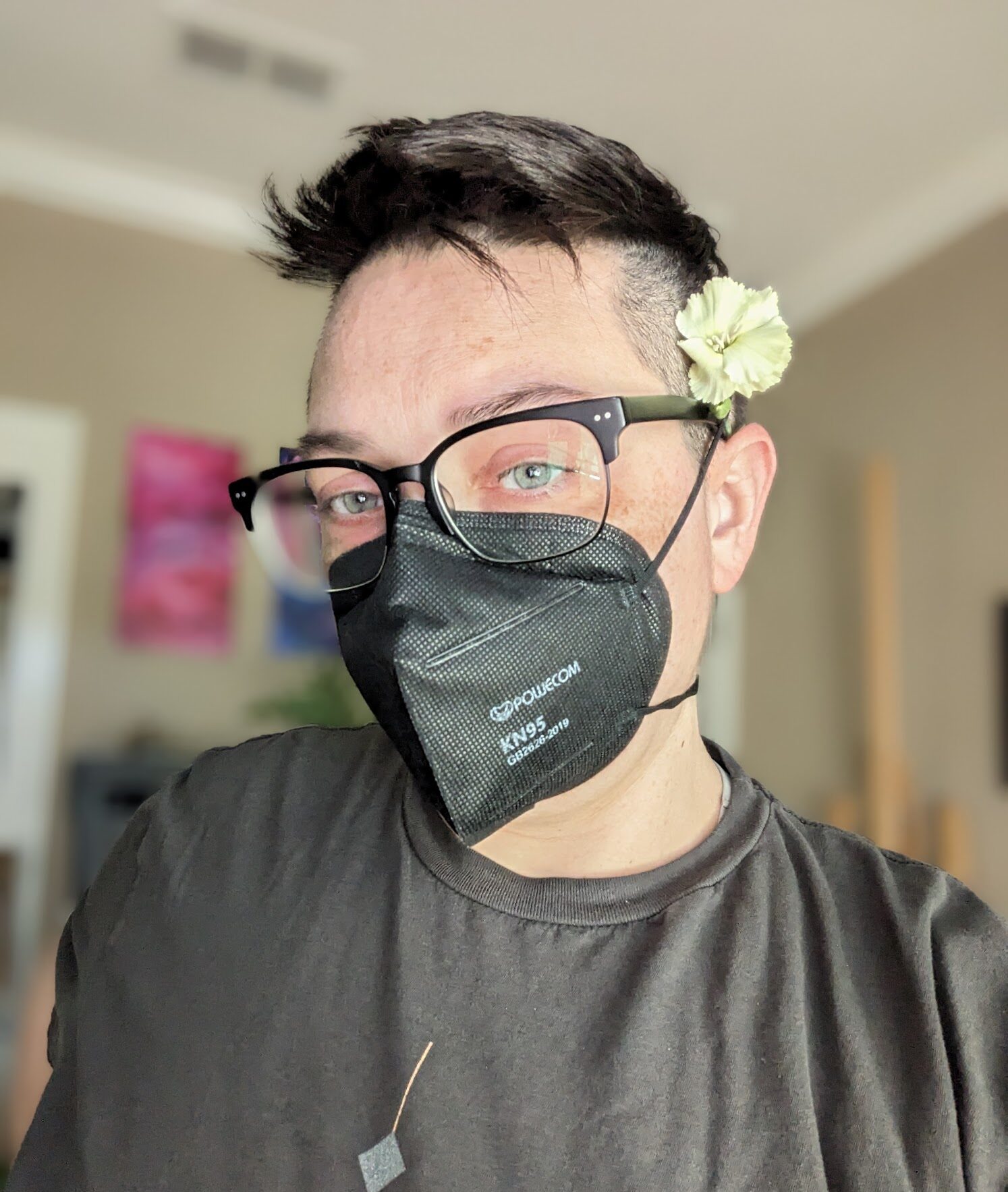 Photo of Emily wearing glasses and a KN95 mask with a green flower behind their ear